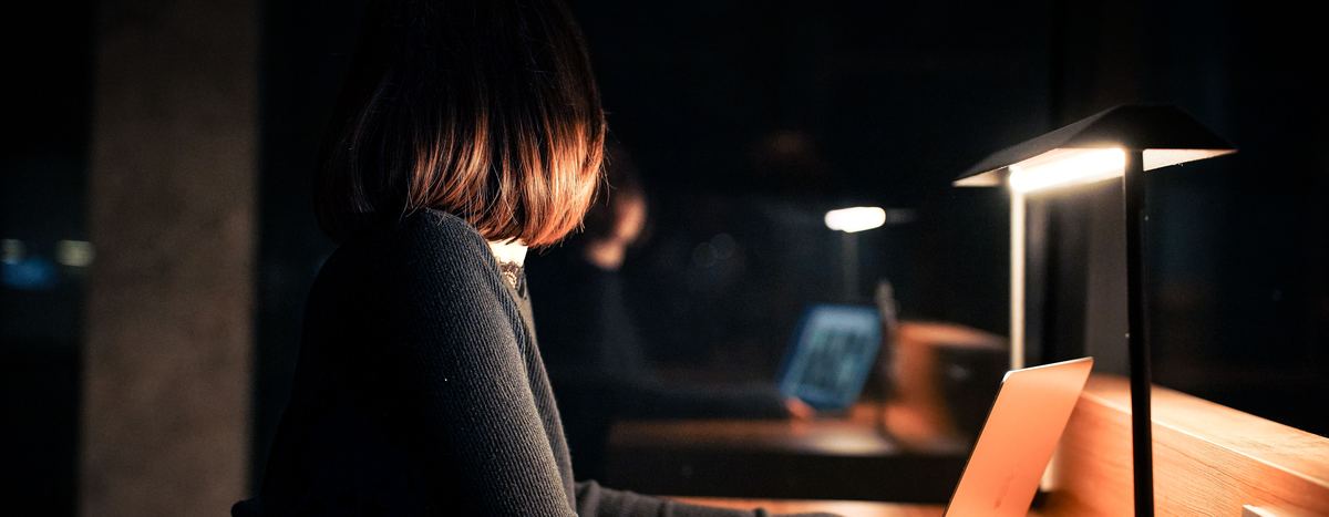 A woman with shoulder-length dark hair sits at a long wooden desk at night, working on an open silver laptop lit by a tall desk lamp.